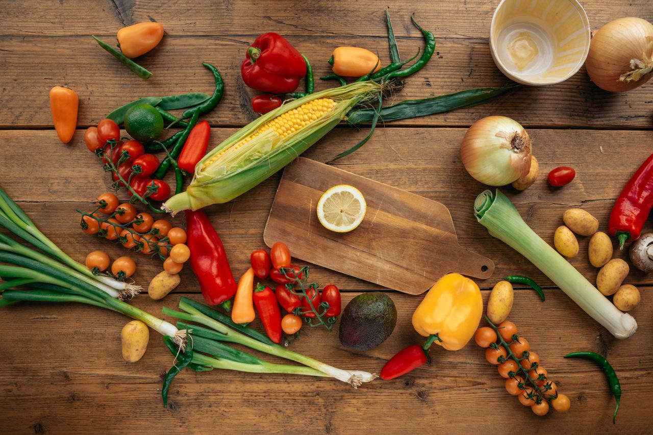 hero-img-01 Colorful assortment of fresh vegetables and fruits arranged on a wooden table with cutting board.